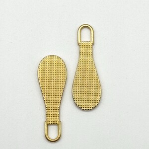 May include: Two gold-colored zipper pulls with a textured, grid-like pattern. The pulls are shaped like teardrops with a loop at the top.