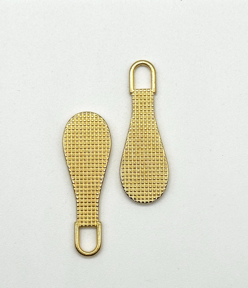 May include: Two gold-colored zipper pulls with a textured, grid-like pattern. The pulls are shaped like teardrops with a loop at the top.