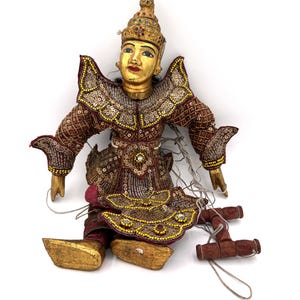 May include: A vintage Burmese marionette doll with a gold painted face and headdress. The doll is dressed in a maroon and gold sequined costume with intricate detailing. The puppet is approximately 12 inches tall.