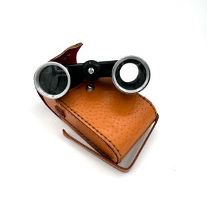 May include: Vintage opera glasses with a black frame and silver-colored lenses, resting on a textured, tan leather case. The case has a small handle and a red interior. The binoculars are a classic accessory.