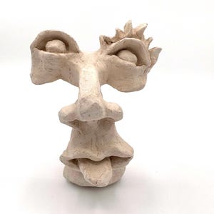 May include: A beige, abstract sculpture of a face with exaggerated features. The sculpture has large, rounded eyes, a prominent nose, and a wide, layered mouth. A crown-like shape sits atop the head. The artwork is set against a white background.