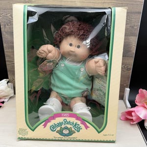 May include: A Cabbage Patch Kids doll with brown yarn hair and a round face, wearing a light green romper and white shoes. The doll is inside a rectangular cardboard box with a clear plastic window, featuring the text 1985 Cabbage Patch Kids.