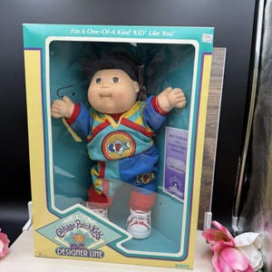 May include: A Cabbage Patch Kids doll in a box with the text I'm A One-Of-A-Kind KID Like You! and Designer Line. The doll has dark hair and wears a blue, red, and yellow outfit with CPK logos. A birth certificate and adoption papers are included.