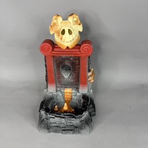May include: A decorative incense burner shaped like a stone fortress with a beige skull at the top. The structure features red pillars, a dark brick-patterned wall with a central face carving, and a black base with a small golden waterfall feature.