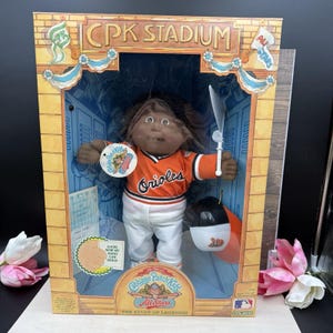 May include: A Cabbage Patch Kids All Stars doll in a CPK Stadium box. The doll has dark skin, brown hair, and wears an orange Orioles jersey with white pants. It holds a white bat and includes a black and white baseball helmet. The box features text.