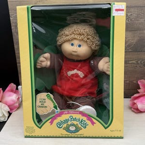 May include: A Cabbage Patch Kids doll with tan yarn hair, blue eyes, and freckles, wearing a red shirt and brown pants. The doll is inside a yellow and green cardboard box with the text I CAN HOLD MY CRAYON and The Official Cabbage Patch Kids.