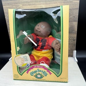 May include: A Cabbage Patch Kids doll in original packaging, featuring a dark-skinned doll wearing a red and yellow jersey with the number 11. The doll holds a small white plastic toothbrush. The box includes text reading The Official Cabbage Patch Kids.
