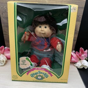 May include: A Cabbage Patch Kids doll with dark brown hair, wearing a red and blue shirt and red pants, packaged in a yellow box. The box features the text I CAN HOLD MY CRAYON and The Official Cabbage Patch Kids, with a small crayon accessory.
