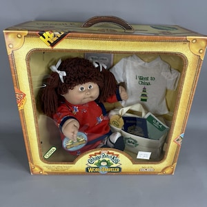 May include: A Cabbage Patch Kids World Traveler doll in a box, featuring brown yarn hair with white bows, a red floral dress, a white t-shirt with the text I Went to China, a miniature passport, and a small beige travel bag with green accents.