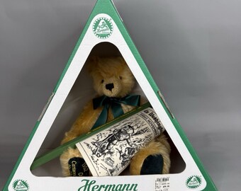 May include: A golden-brown plush teddy bear with a dark green ribbon around its neck, seated inside a triangular green cardboard display box. The box features the Hermann logo and a printed replica of a 1902 Washington newspaper front page.