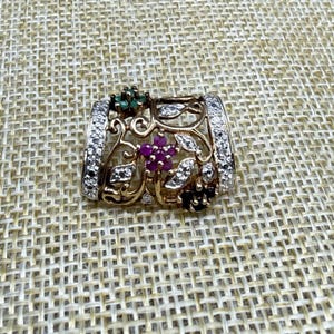 May include: A gold-toned ring with a floral design. The ring features a central cluster of small, round, red gemstones, surrounded by white gemstones and green and black accents. The band is encrusted with white gemstones.