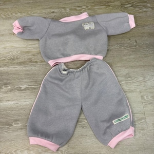 May include: A gray two-piece outfit consisting of a long-sleeved pullover top and matching pants. The top features light pink trim at the collar and cuffs, with a small white cat patch on the chest. The pants have light pink cuffs and a small rectangular label.