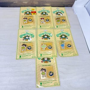 May include: Seven Cabbage Patch Kids pin and ring sets on individual yellow cardboard backing cards. Each set includes a small character figure pin and a matching ring featuring a cabbage leaf design, all encased in clear plastic blister packs.