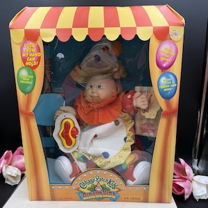 May include: A Cabbage Patch Kids Circus Kids doll in a box with a red and yellow striped top. The doll wears a clown outfit with a ruffled collar and holds a red and yellow accessory. The packaging features text including Look! Now my hand can hold!, Funny-face mask included!, Crazy clown shoe that squeaks!, and Bonus poster & trading card inside!