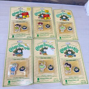 May include: Six Cabbage Patch Kids pin and ring sets in original packaging. Each set includes a small character figure pin and a cabbage-shaped ring. The cards feature green text, a cabbage logo, and the Howard Eldon, Ltd. brand name.