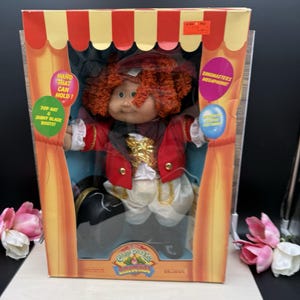 May include: A Cabbage Patch Kids doll dressed as a ringmaster, featuring curly red hair, a red jacket with gold accents, white pants, and black boots. The doll is packaged in a box with circus-themed graphics, including text like Hand That Can Hold.