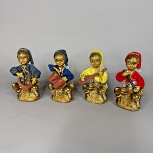 May include: Four figurines of musicians sitting on gold-colored bases. Each figure wears a hat and a fabric shirt in blue, dark blue, yellow, or red. They hold musical instruments including a lyre, drums, a lute, and a flute, with pearl accents.