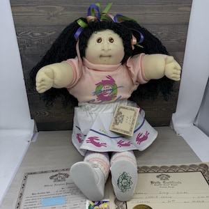 May include: A Cabbage Patch Kids doll with black yarn hair, brown eyes, and a peach-colored shirt featuring a rabbit graphic. The doll wears a white skirt with purple rabbit prints, white socks, and white shoes. A tag reads Lily Lilac.