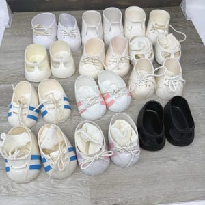 May include: A collection of twenty-two doll shoes in various styles, including white sneakers with blue or pink stripes, solid white lace-up shoes, and a pair of solid black shoes. Several shoes contain small white fabric socks inside the footwear.