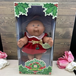May include: A Cabbage Patch Kids Holiday Baby doll in a box. The doll has a round face, light skin, and brown eyes. It wears a red dress with a white lace collar, a gold bow, and a holly graphic. The box features holly leaves and berries.
