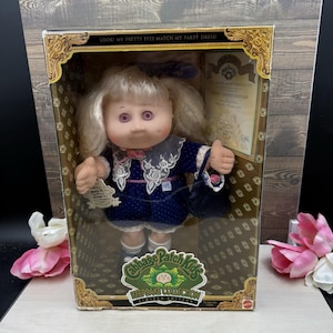 May include: A Cabbage Patch Kids doll from the Keepsake Collection in a display box. The doll has blonde hair, purple eyes, and wears a navy blue dress with white lace trim and a matching purse. The box text reads Look! My pretty eyes match my party dress!