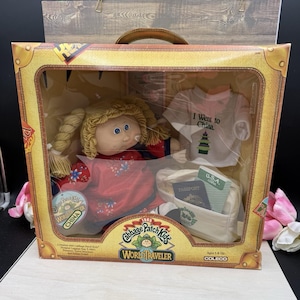May include: A Cabbage Patch Kids World Traveler doll set in a box featuring a blonde doll with braided hair in a red dress, a white t-shirt with the text I Went to China, a small white bag, a passport, and a travel brochure with U.S.A. China text.