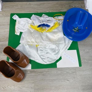 May include: A white satin shirt with yellow fringe trim and a blue collar, a bright blue plastic cowboy hat with a gold star emblem, and a pair of brown plastic boots. The set includes a white plastic tag and a white plastic strap component.