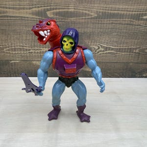 May include: A Skeletor action figure with light blue skin, a yellow skull face, and a purple hood. The figure wears a purple and red chest harness with a red dragon head attachment over the right shoulder and holds a purple sword in the right hand.