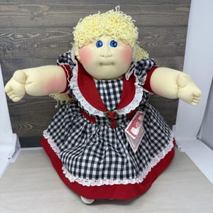 May include: A soft-sculpture doll with blonde yarn hair, blue eyes, and rosy cheeks. The doll wears a red and black gingham dress featuring a white eyelet lace collar, red trim, and two small red fabric roses at the bodice. A white tag is attached.
