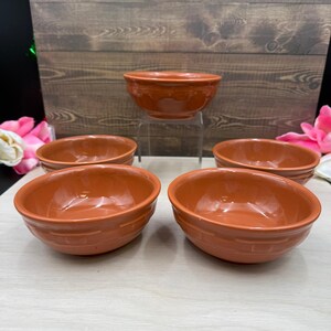 May include: A set of five terracotta-colored ceramic bowls with a smooth, glossy finish. Each bowl features a subtle, embossed grid pattern around the exterior base. The bowls are arranged with one elevated on a clear stand and four positioned below.