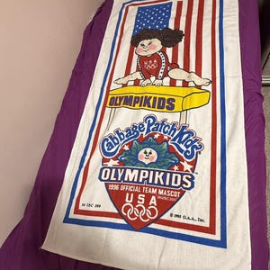 May include: A white rectangular beach towel featuring a Cabbage Patch Kids character performing gymnastics on a yellow pommel horse. The design includes red and blue stripes, the text OLYMPIKIDS, 1996 OFFICIAL TEAM MASCOT, USA, and Olympic rings.