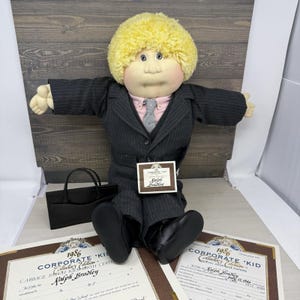 May include: A Cabbage Patch Kids doll with yellow yarn hair, wearing a dark gray pinstripe suit, a light pink collared shirt, and a gray tie. The doll has black shoes, a black briefcase, and two 1986 Corporate Kid Collector's Edition documents.