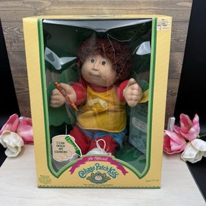 May include: A Cabbage Patch Kids doll with brown yarn hair, wearing a yellow shirt and red pants, packaged in a yellow box. The box features the text 'I CAN HOLD MY CRAYON!' and 'The Official Cabbage Patch Kids' with a small orange crayon accessory.