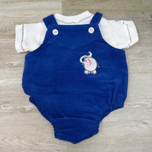 May include: A royal blue corduroy overall romper featuring two white buttons on the straps and an embroidered elephant patch with a pink ear. The romper is layered over a white short-sleeved shirt with a subtle dark blue stitched trim.