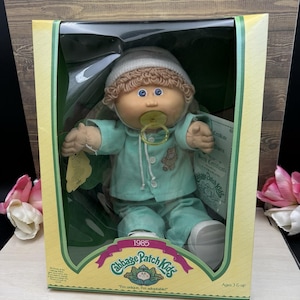 May include: A Cabbage Patch Kids doll from 1985 in its original packaging. The doll has light brown yarn hair, a white knit cap, and a light green pajama set with a small bear graphic. A yellow pacifier is in its mouth. The box includes a birth certificate.