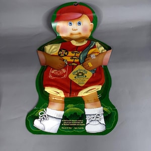 May include: A Cabbage Patch Kids doll romper in red and yellow, packaged on a green cardboard display shaped like a child. The outfit features a firefighter-style design with a badge graphic, white shoes, and text stating it fits 16 inch kids.