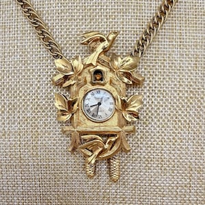 May include: Gold-toned necklace featuring a cuckoo clock pendant. The pendant has a clock face with Roman numerals, surrounded by leaves, birds, and pinecone details. The chain is a thick, textured link.
