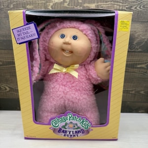 May include: A Cabbage Patch Kids Babyland Bunny doll in a pink fuzzy hooded suit with long ears, blue eyes, and a single tooth. The doll wears a yellow ribbon at the neck. The box features the text SQUEEZE MY TAIL - IT SQUEAKS! and Babyland Bunny.