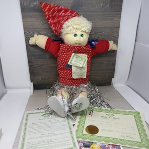 May include: A soft-sculpture doll with blonde yarn hair, blue eyes, and a red hat. The doll wears a red long-sleeved shirt under a red and white polka-dot jumper with a silver tinsel skirt. It includes a name tag and two official documents.