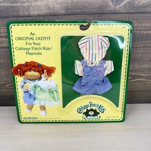 May include: A Cabbage Patch Kids Playmate original outfit set in its original packaging. The set includes a white shirt with thin multicolored stripes, a matching cap, and blue denim-style overalls. The packaging features a doll illustration.