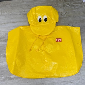 May include: A bright yellow rain poncho featuring a hood with two white and black cartoon eyes on the front. The garment has a smooth, glossy texture and includes a small red rectangular patch on the chest with the white letters CPK printed on it.