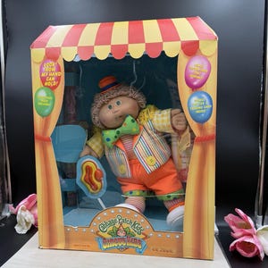 May include: A Cabbage Patch Kids Circus Kids doll in a box, wearing a striped vest, orange pants, and a green polka-dot bow tie. The doll has brown yarn hair and a small hat. The packaging features red and yellow stripes and text including Look! Now My Hand Can Hold! and Bonus Poster & Trading Card Inside!