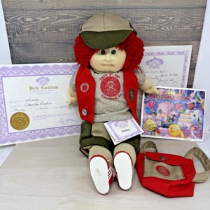 May include: A Cabbage Patch Kids doll with red yarn hair wearing a tan cap, a gray t-shirt with red Camp Cabbage text, a red vest, and tan shorts. The doll includes a birth certificate, adoption papers, a birthday card, and a red and tan backpack.