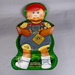 May include: A Cabbage Patch Kids doll romper in blue denim with yellow trim, packaged on a green cardboard display featuring a cartoon character wearing a red cap, yellow shirt, and white shoes, with text including "Cabbage Patch Kids" and "ROMPER".