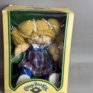 May include: A Cabbage Patch Kids doll with blonde yarn hair in pigtails, blue eyes, and a plaid dress with a blue ribbon. The doll wears blue leggings and white shoes, packaged in a yellow box with the Cabbage Patch Kids logo printed on it.