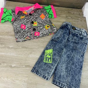 May include: A short-sleeve button-down shirt with a black and white speckled pattern, neon pink collar and sleeve cuffs, and neon orange and green diamond accents. Paired with acid-wash blue denim jeans featuring a neon green rectangular patch.