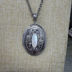 May include: Silver-toned oval locket necklace with an ornate floral design and a pearlescent oval center. The locket hangs from a matching chain. The background is a neutral tan fabric.