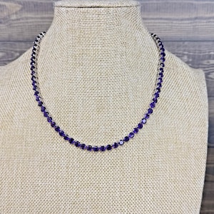 May include: A necklace with small, round, deep purple gemstones. The gemstones are strung together with a silver-colored chain. The necklace is displayed on a beige mannequin.
