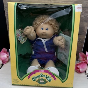 May include: A Cabbage Patch Kids doll from 1985 with curly blonde hair and blue eyes, wearing a dark blue short-sleeved outfit with a white collar. The doll is packaged in a yellow and green cardboard box with clear plastic front window.