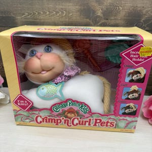 May include: A Cabbage Patch Kids Crimp 'n Curl Pets kitty toy in its original yellow and pink box. The white plush cat features blue eyes, a pink nose, a pink patterned collar, and a blue fish tag. The packaging includes text and images.